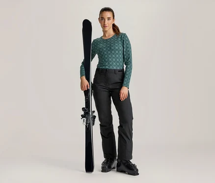 Femme debout, tenant un ski noir. Elle porte un haut à manches longues vert à motifs, un pantalon de ski noir et des chaussures de ski noires.
