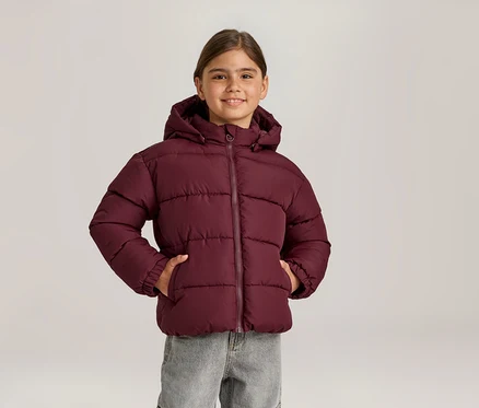 Jeune fille souriante portant une doudoune bordeaux à capuche et un jean gris, les mains dans les poches de sa veste.