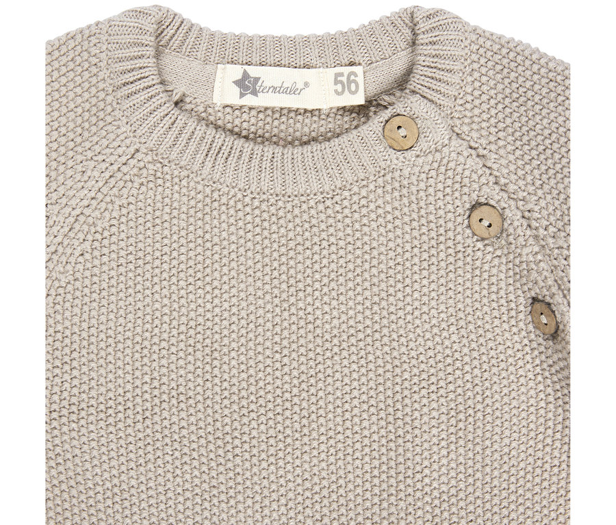 Gros plan d'un pull bébé tricoté beige clair avec un col côtelé, trois boutons en bois sur l'épaule droite et une étiquette blanche "Sterntaler 56" à l'arrière du cou.