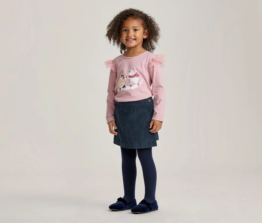 Jeune fille aux cheveux bouclés, souriante, portant un haut rose à manches longues avec un motif d'ours polaires et des volants aux épaules, une jupe bleu foncé, des collants bleu foncé et des chaussures en velours bleu foncé.