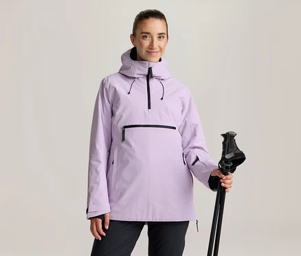 Femme portant une veste anorak violet clair avec des fermetures éclair noires et un pantalon noir, tenant des bâtons de ski noirs.