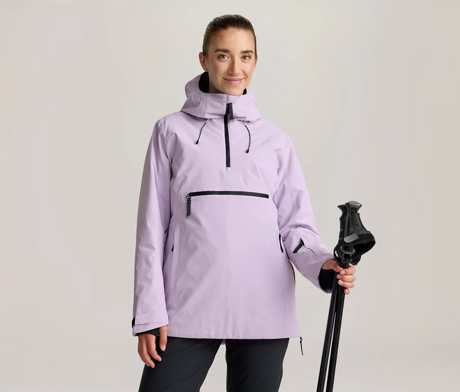 Femme portant une veste anorak violet clair avec des fermetures éclair noires et un pantalon noir, tenant des bâtons de ski noirs.
