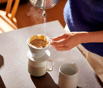 Une personne verse de l'eau chaude d'une bouilloire dans un filtre à café manuel blanc, taille 101, posé sur une tasse. De la vapeur s'échappe du filtre. Une autre tasse est sur la table.