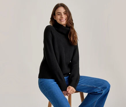 Femme souriante aux longs cheveux bruns, portant un pull à col roulé noir et un jean bleu, assise sur un tabouret en bois.