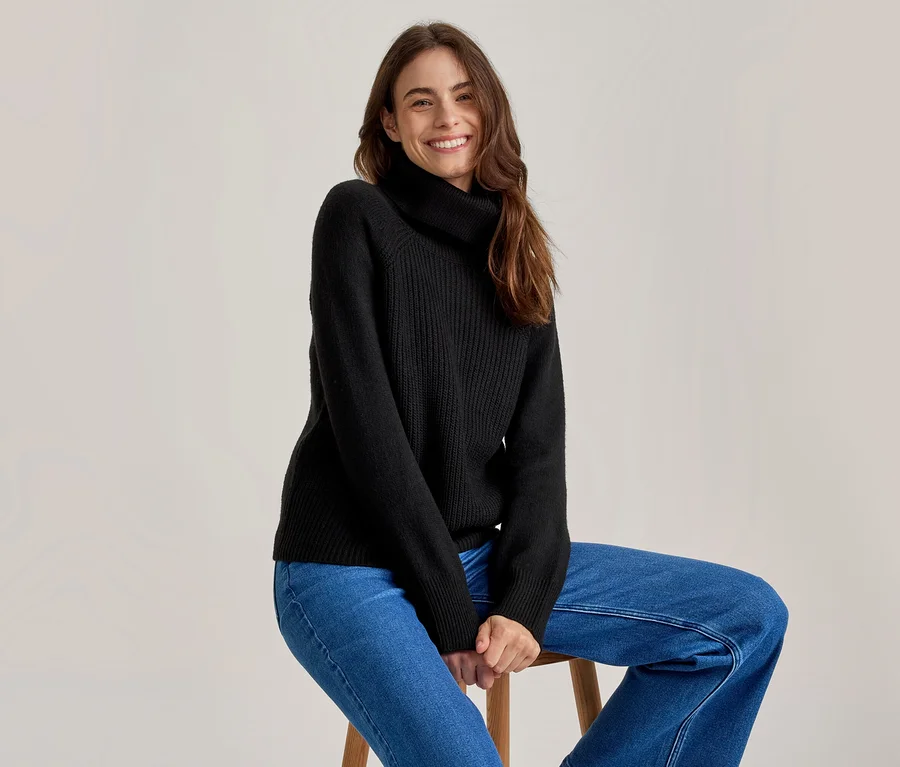 Femme souriante aux longs cheveux bruns, portant un pull à col roulé noir et un jean bleu, assise sur un tabouret en bois.