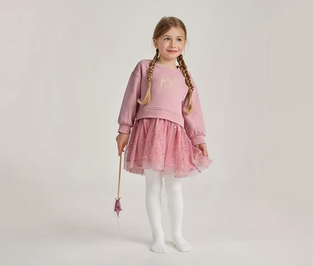 Une jeune fille aux tresses blondes porte un haut rose à manches longues avec une étoile dorée, une jupe tutu rose pailletée et des collants blancs, tenant une baguette étoilée et souriant.