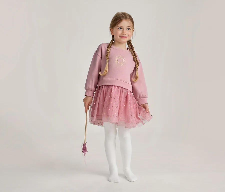 Une jeune fille aux tresses blondes porte un haut rose à manches longues avec une étoile dorée, une jupe tutu rose pailletée et des collants blancs, tenant une baguette étoilée et souriant.