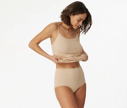 Une femme aux cheveux courts et bruns porte un débardeur beige à fines bretelles et une culotte taille haute assortie. Elle regarde vers le bas et remonte l'ourlet de son débardeur avec les deux mains.