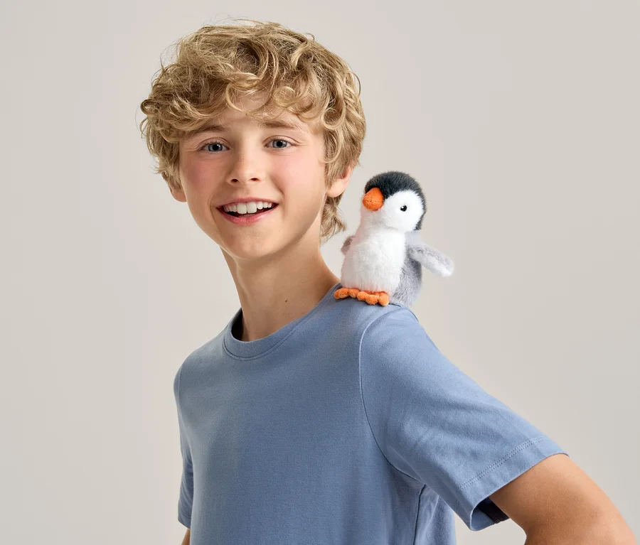 Garçon souriant aux cheveux blonds bouclés portant un t-shirt bleu, un petit pingouin en peluche est assis sur son épaule droite.