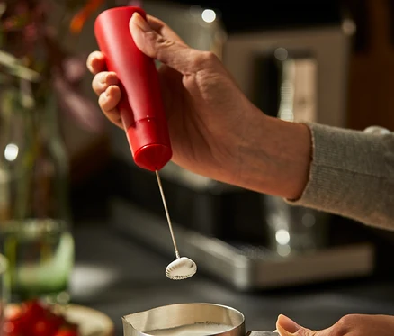 Une femme utilise un mousseur à lait rouge à piles pour faire mousser du lait dans un pot à lait en acier inoxydable.