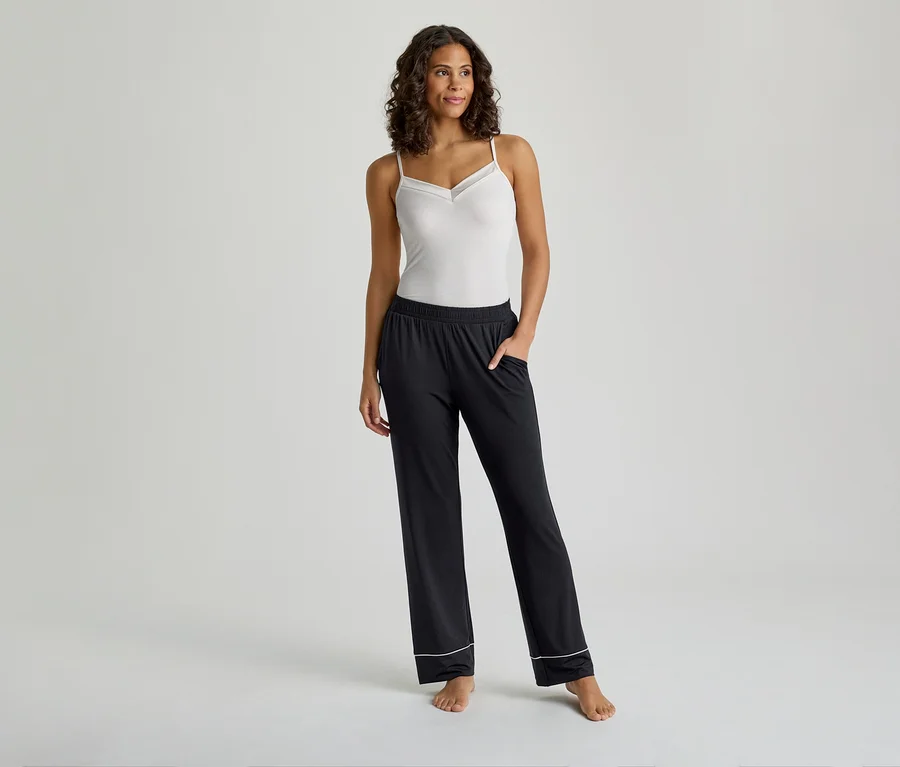 Une femme se tient de face, vêtue d'un caraco blanc avec une bordure délicate et d'un pantalon de pyjama noir avec un passepoil blanc aux poignets. Ses mains sont dans ses poches.