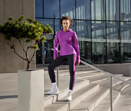 Femme en veste technique pour femme FILA violette et leggings noirs, posant sur des marches avec un pied sur un bloc.
