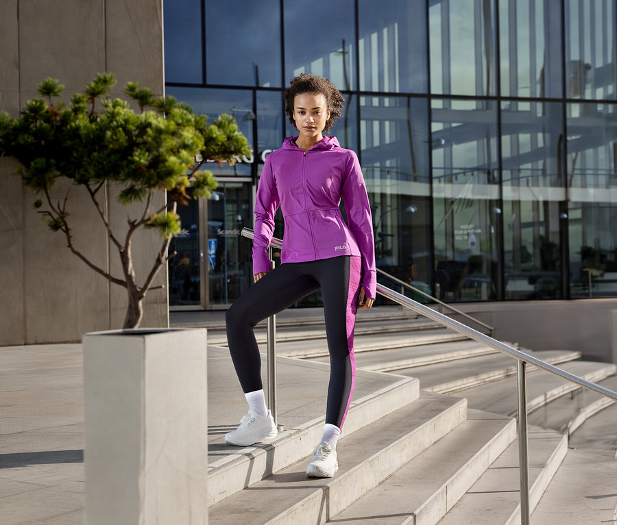Femme en veste technique pour femme FILA violette et leggings noirs, posant sur des marches avec un pied sur un bloc.
