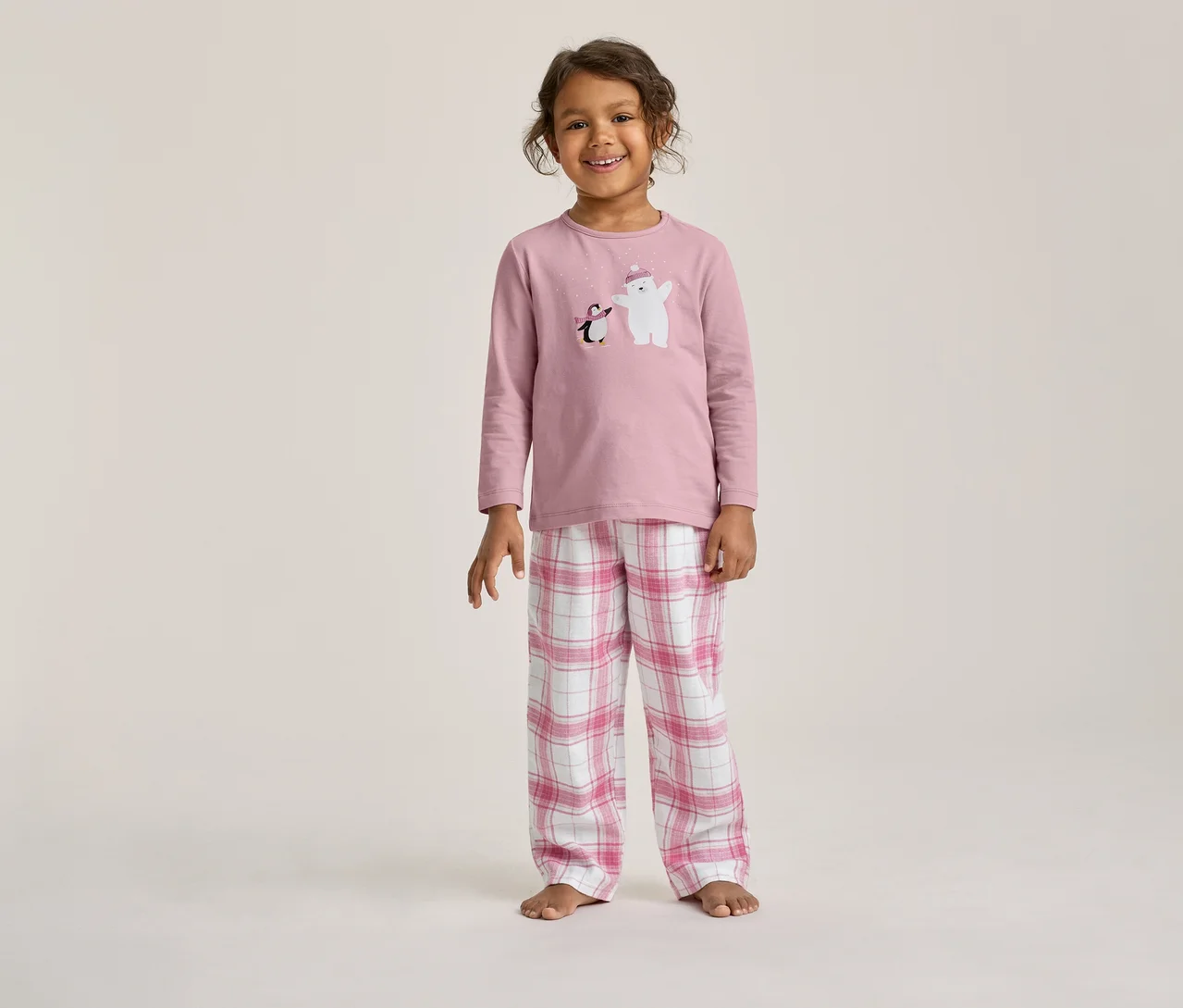 Enfant souriant portant un pyjama rose à manches longues avec un imprimé ours polaire et pingouin, et un pantalon à carreaux roses et blancs.