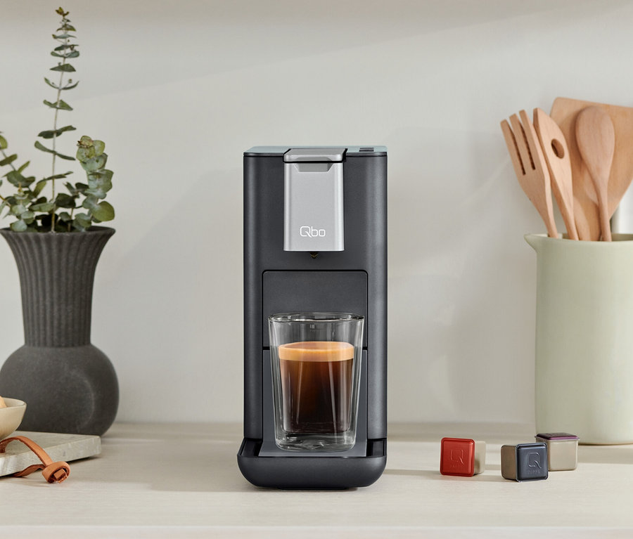 Une machine à café Qbo ESSENTIAL Dark Stone avec une tasse de café. À côté, des capsules et un bol de framboises.