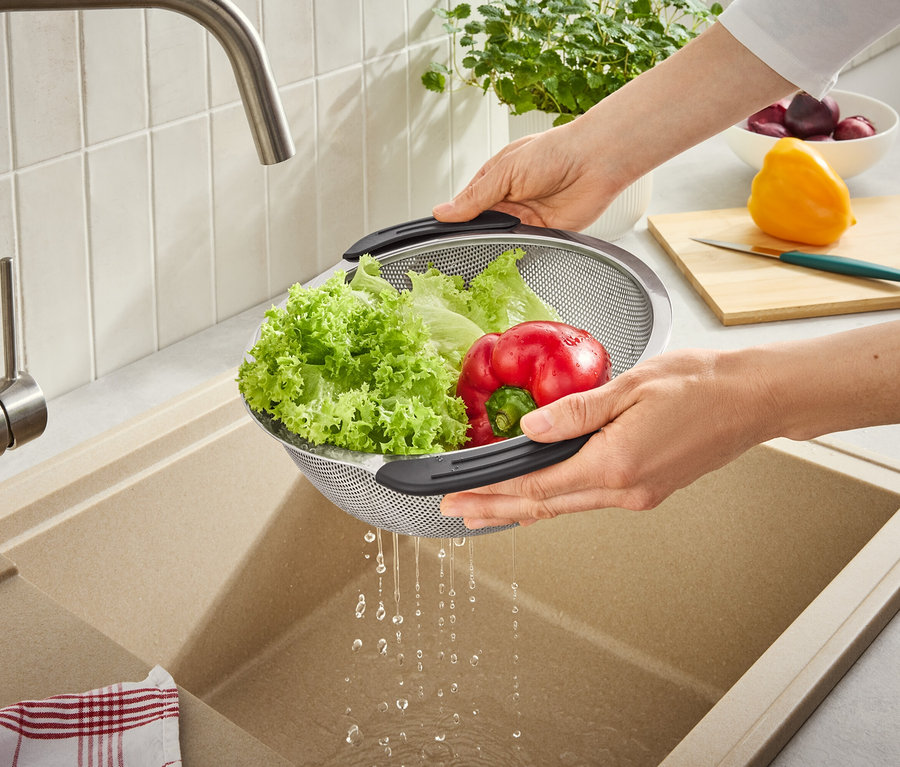 Des mains rincent de la laitue verte et un poivron rouge dans une passoire métallique sous un robinet, l'eau s'écoulant dans un évier de cuisine. Une planche à découper et d'autres légumes sont en arrière-plan.