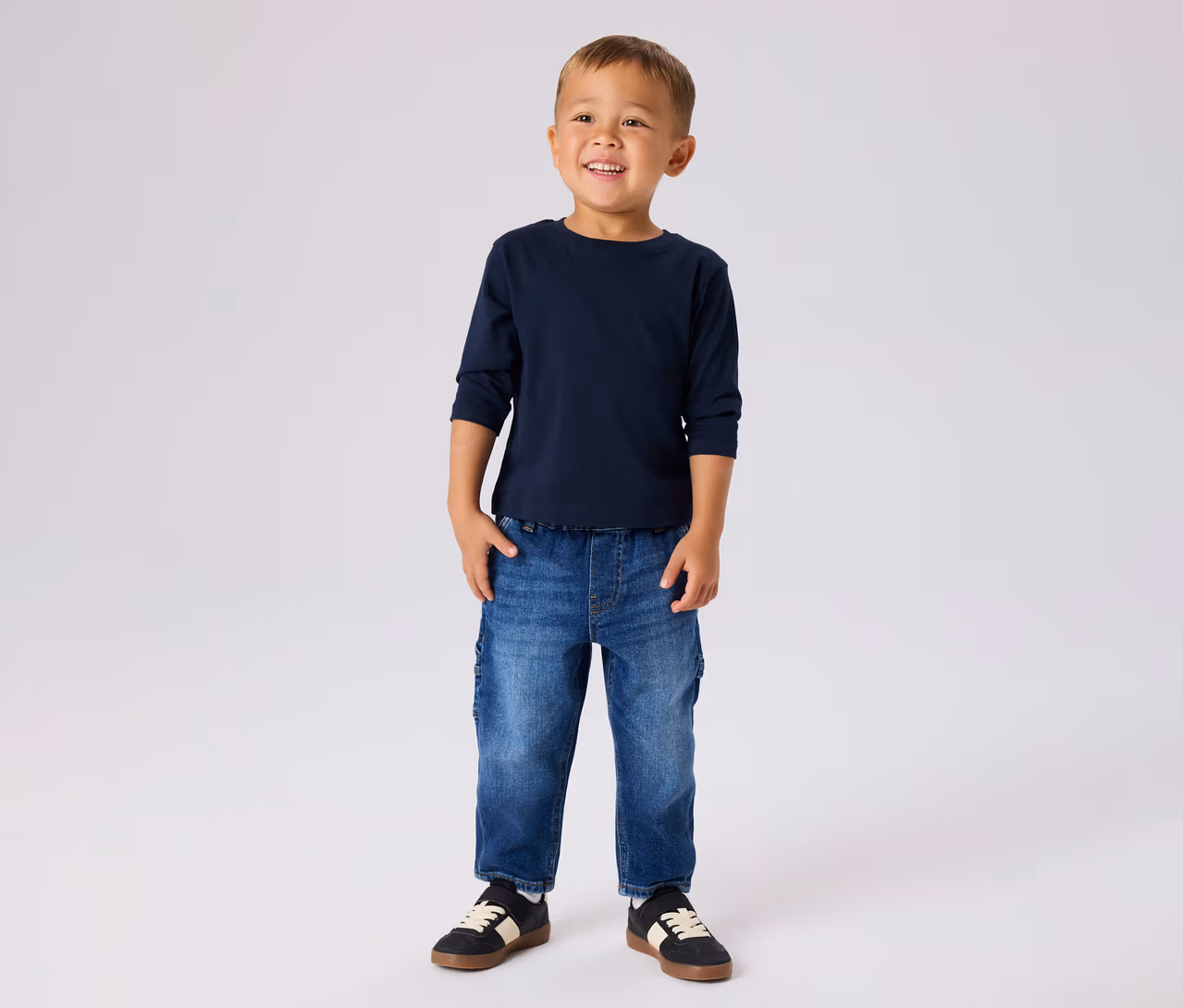 Jeune garçon souriant, portant un t-shirt bleu marine à manches longues, un jean bleu et des baskets noires à rayures blanches.