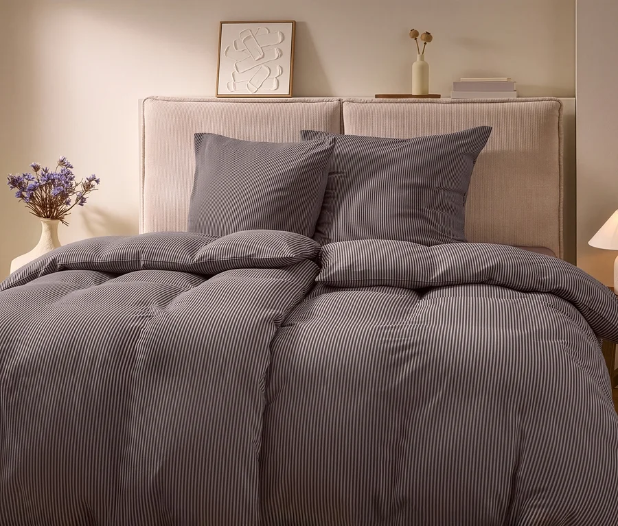 Un lit avec une parure de lit en jersey hivernal rayée, deux oreillers et une tête de lit claire. À gauche, un vase de fleurs séchées, à droite, une lampe sur une table en bois.