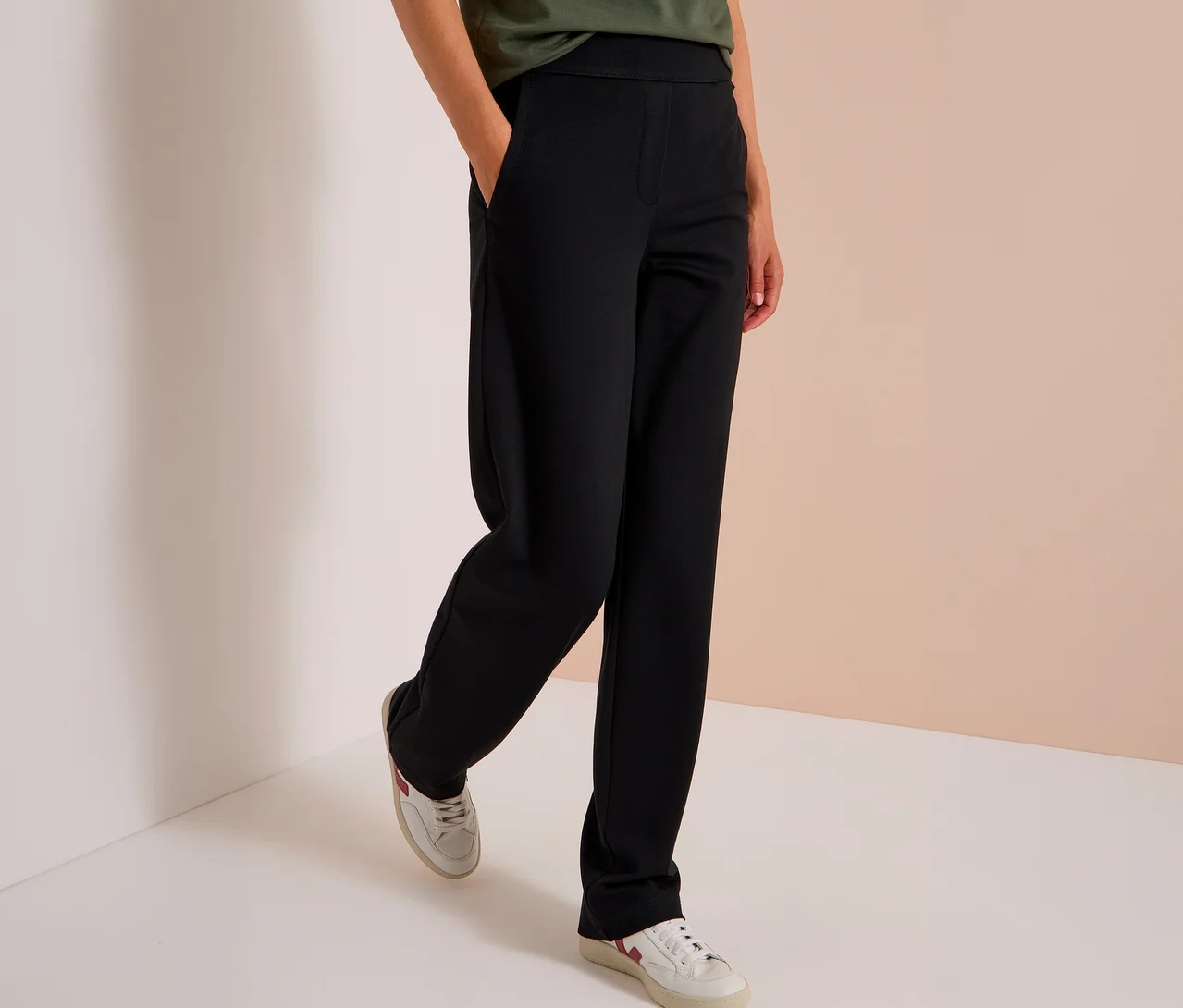 Personne debout, portant un pantalon en tissu CECIL noir et des baskets blanches, une main dans la poche du pantalon.