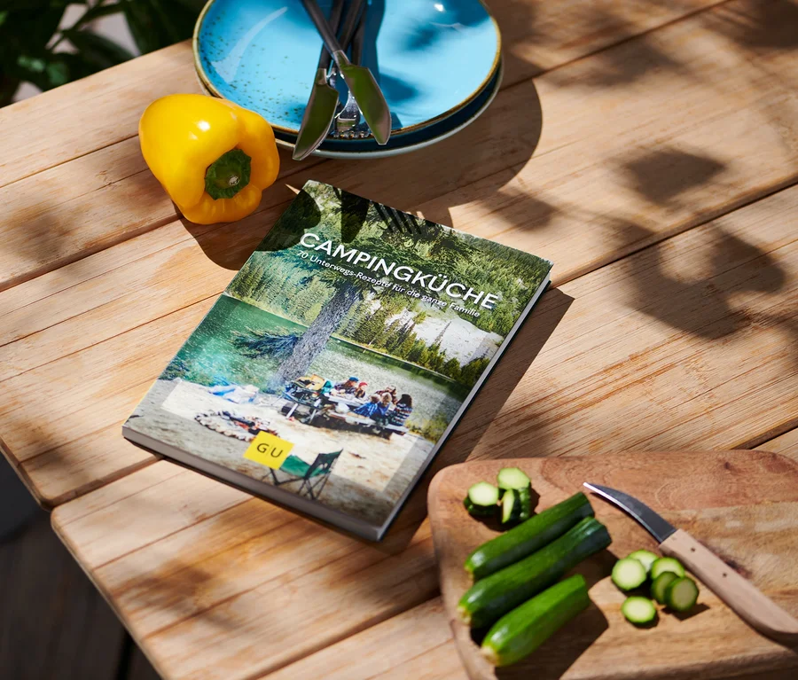 Sur une table en bois se trouvent le livre « Campingküche » (en allemand), un poivron jaune, des assiettes bleues avec des couverts et des courgettes coupées avec un couteau.