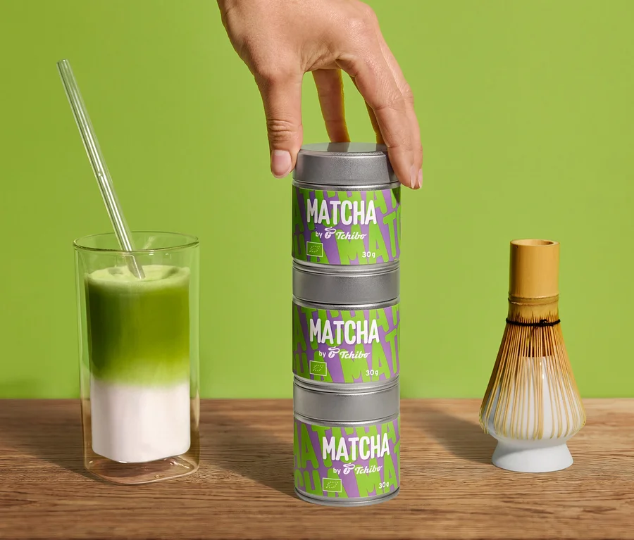 Main empile trois boîtes Bio Matcha by Tchibo - 30 g. Matcha latte en verre avec paille et fouet à matcha sur bois devant vert.