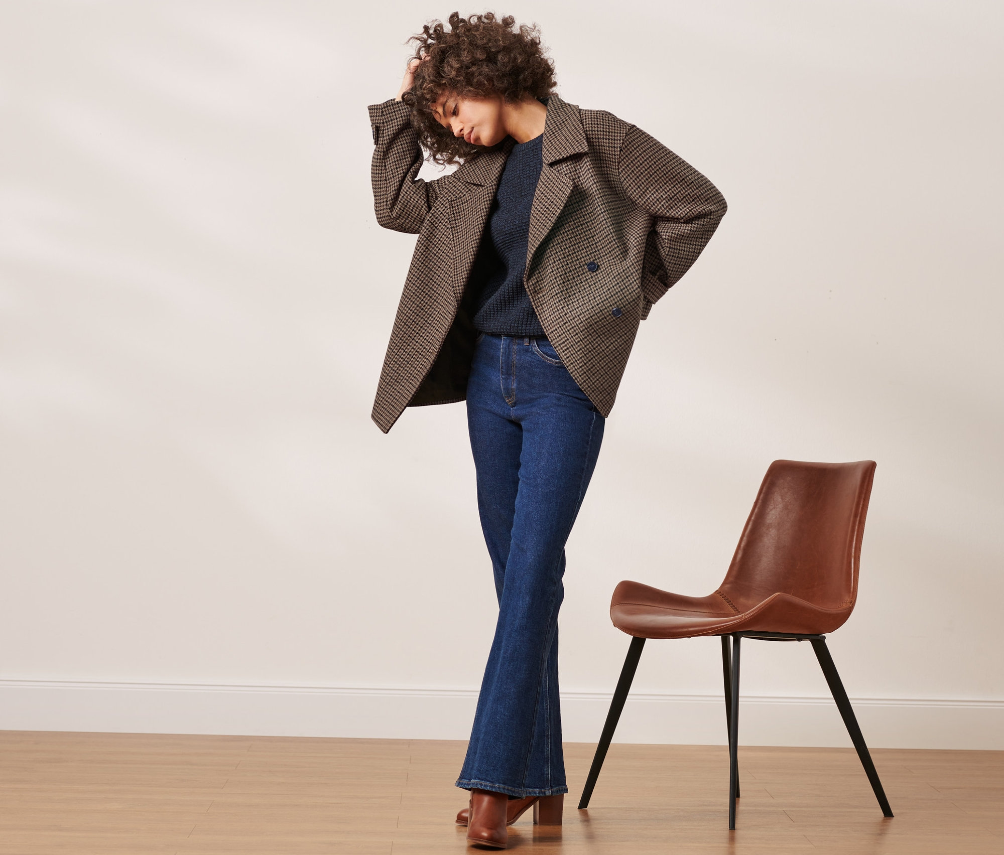 Femme posant en veste à carreaux avec de la laine, pull, jean et chaussures à côté d'une chaise marron.