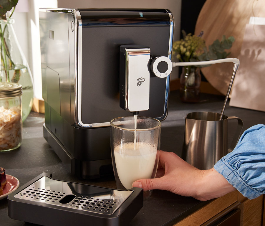 Une femme verse du lait d'une machine à café entièrement automatique Tchibo « Esperto Pro », anthracite, dans un verre. Sur la table, il y a aussi des machines à café Tchibo « Esperto Pro » Dark Red et Metallic Mint.