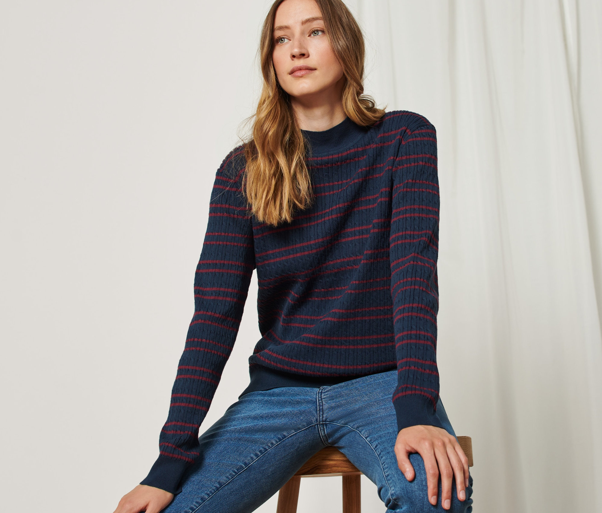 Femme assise sur un tabouret portant un pull en maille fine bleu rouge avec torsades.
