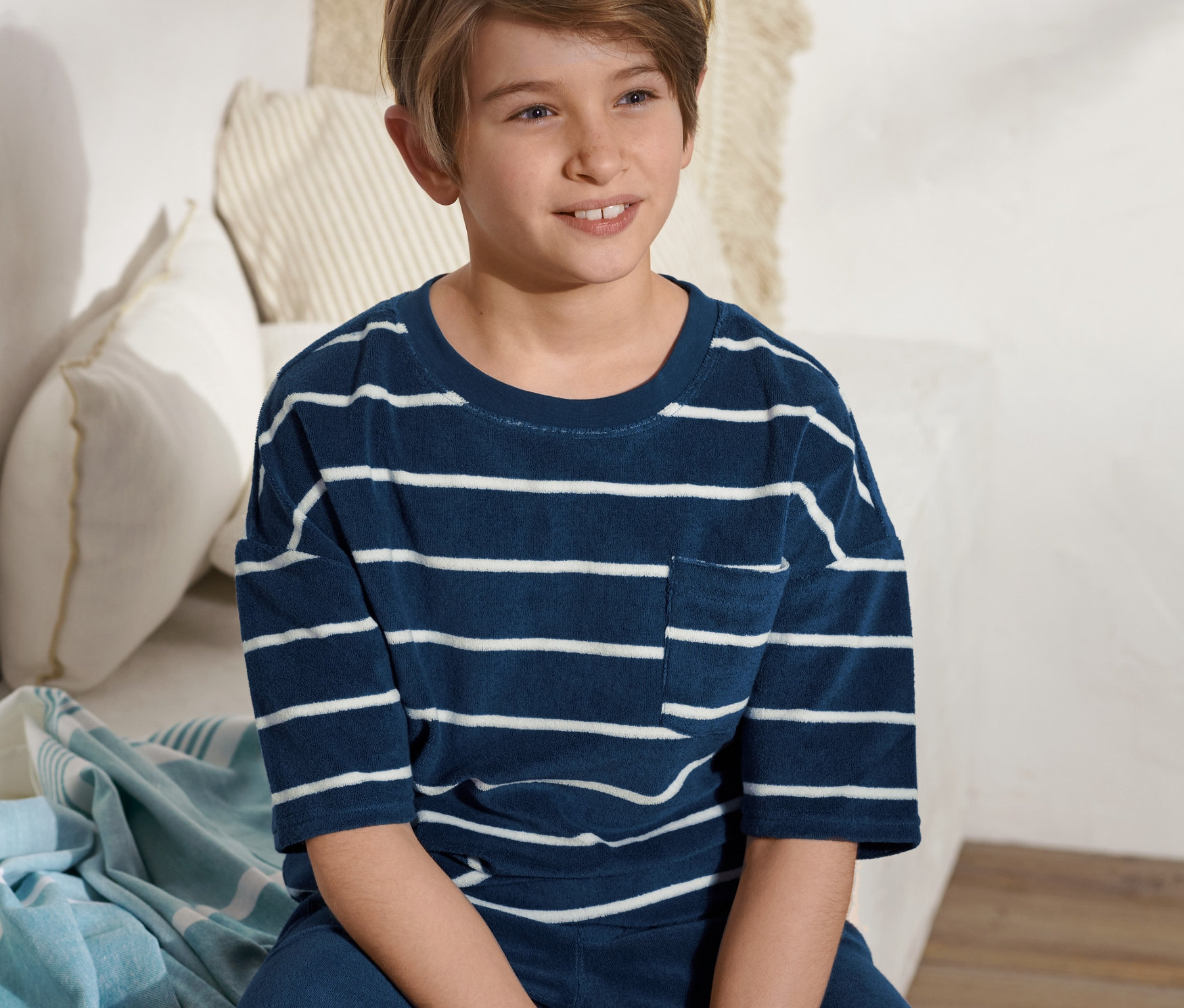 Un garçon est assis sur un lit et porte un t-shirt en éponge bleu et blanc rayé.