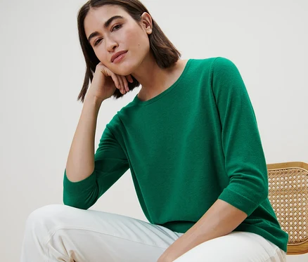 Une femme est assise sur une chaise et porte un pull vert en maille fine avec de la soie et un pantalon blanc.