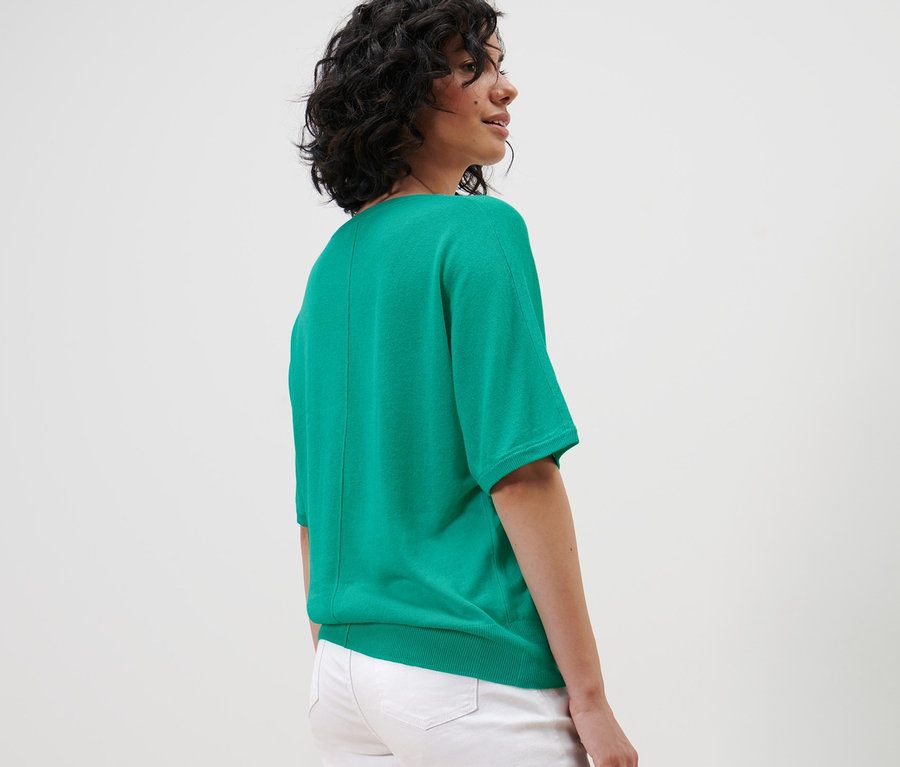 Une femme aux cheveux courts et ondulés pose dans un haut à maille fine avec du lin et un jean blanc.