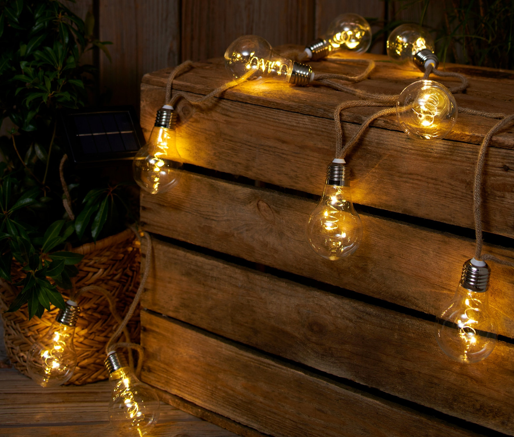 Une guirlande lumineuse solaire avec cordon en jute est posée sur une caisse en bois à côté d'une plante.