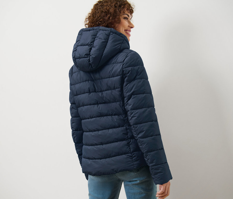 Femme aux cheveux bouclés regardant par-dessus son épaule portant une veste matelassée à capuche bleu foncé.