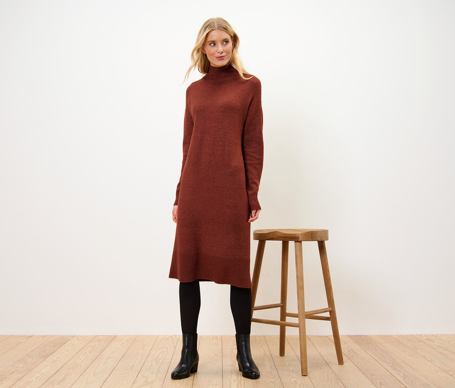 Femme debout à côté d'un tabouret en bois portant une robe en maille avec teneur en laine.