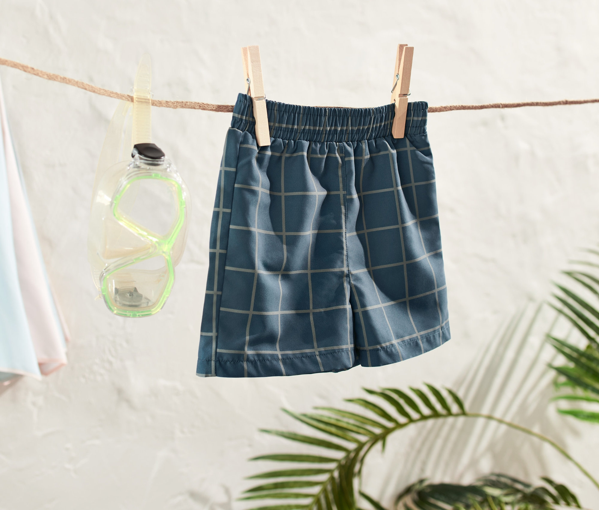 Un boardshort pour tout-petit est suspendu à une corde à linge, à côté de lunettes de plongée et d'un parasol de plage.