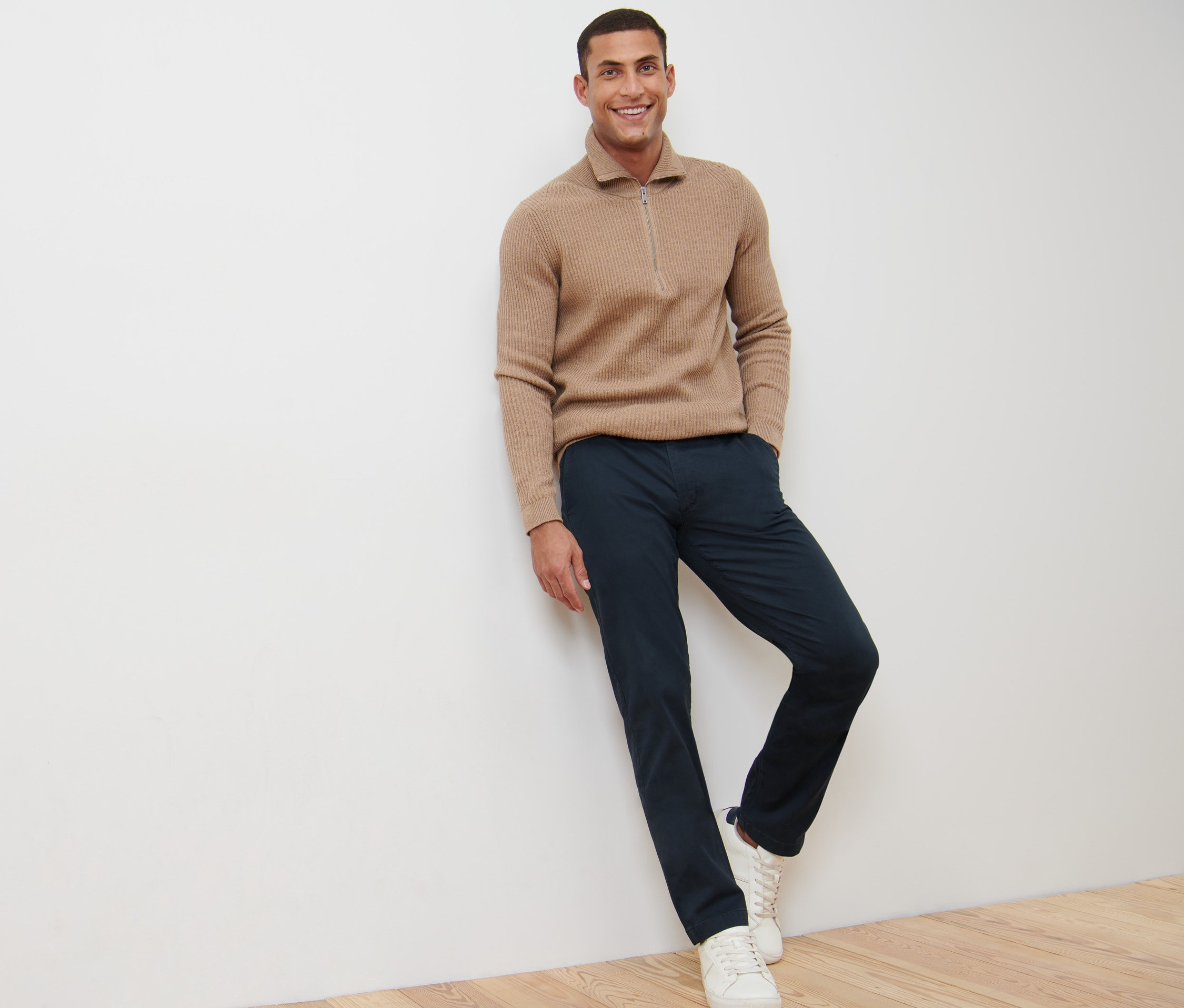 Un homme souriant s'appuie contre un mur et porte un pantalon chino bleu marine.