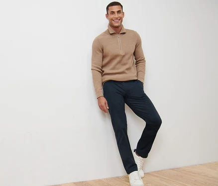 Un homme souriant s'appuie contre un mur et porte un pantalon chino bleu marine.