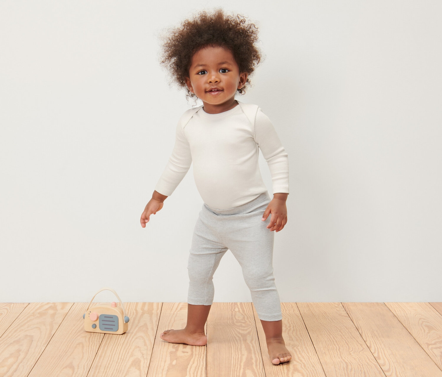Un enfant debout sur un plancher en bois porte 3 bodys côtelés manches longues pour bébé et un legging gris.