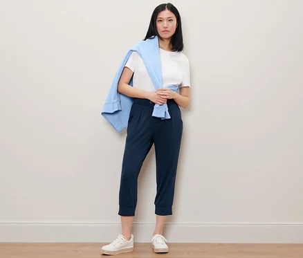 Une femme debout portant un pantalon 3/4 en jersey, un t-shirt blanc et un pull bleu sur les épaules.