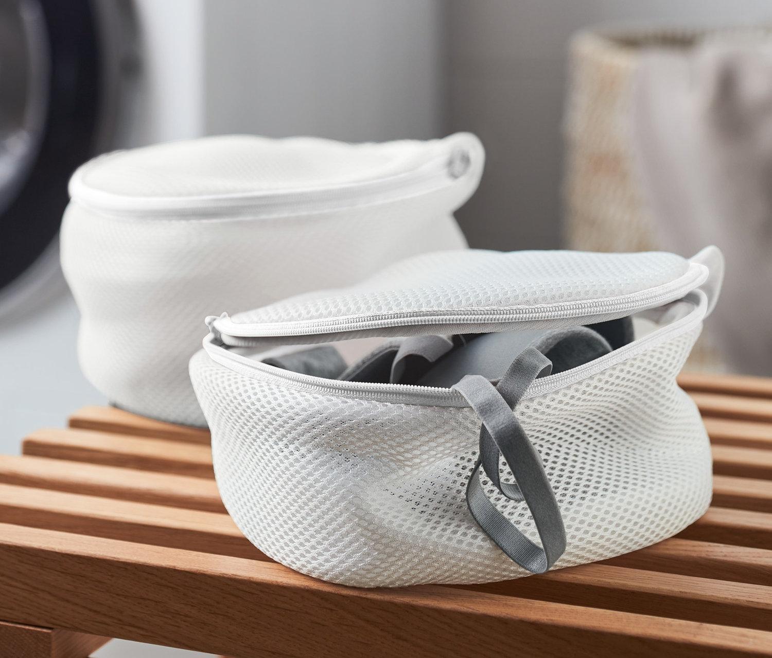 Deux filets de lavage pour soutiens-gorge blancs sur un banc en bois, avec une machine à laver en arrière-plan.