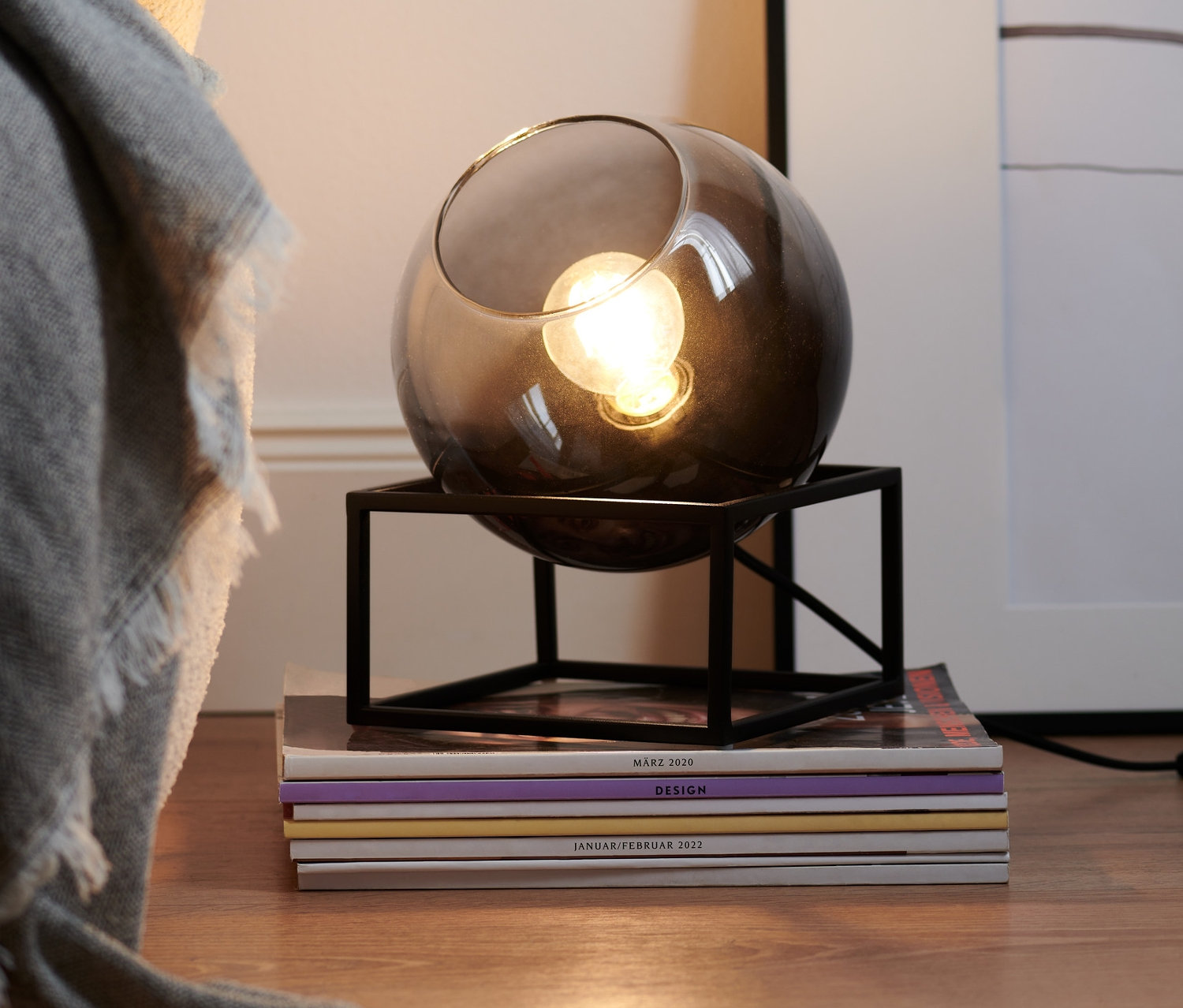 Lampe de table avec abat-jour en verre fumé posée sur une pile de magazines à côté d'une couverture grise.