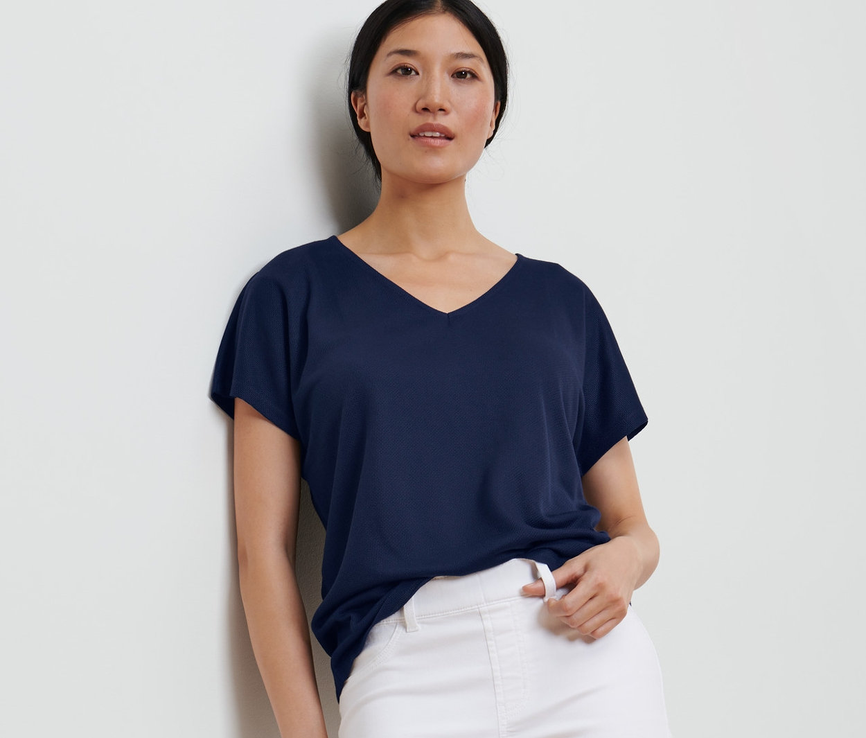 Une femme appuyée contre un mur porte une blouse structurée bleu foncé et un pantalon blanc.