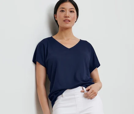 Une femme appuyée contre un mur porte une blouse structurée bleu foncé et un pantalon blanc.