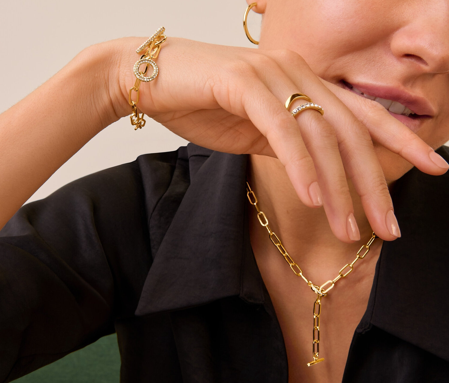 Une femme portant une bague double vague avec cristaux, un bracelet et une chaîne Toggle avec insert de cristaux et des créoles dorées.