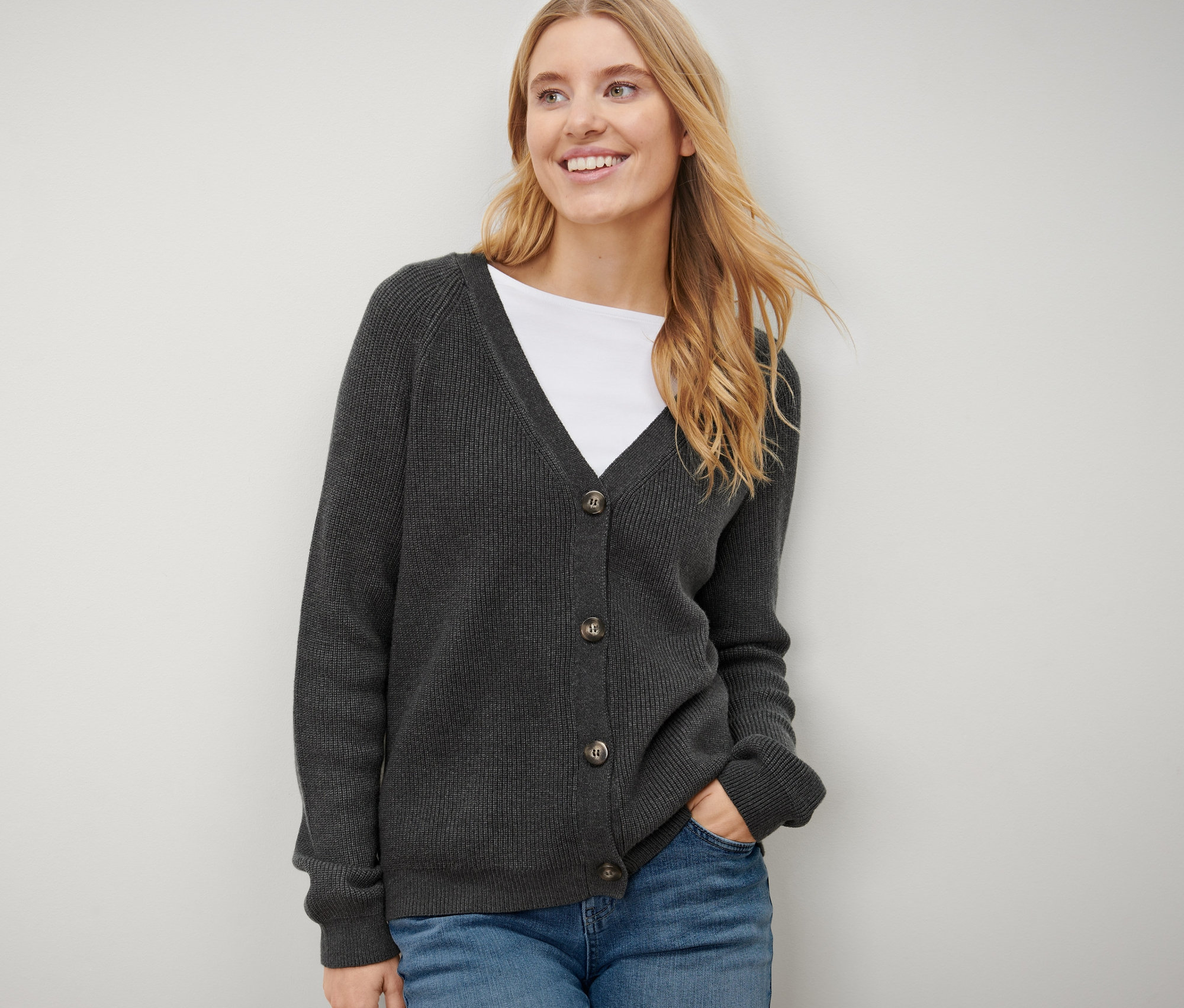 Une femme blonde pose en jean bleu et porte un cardigan en grosse maille.