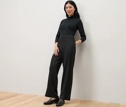 Femme portant un col roulé noir et un pantalon en jersey confortable, appuyée contre le mur.
