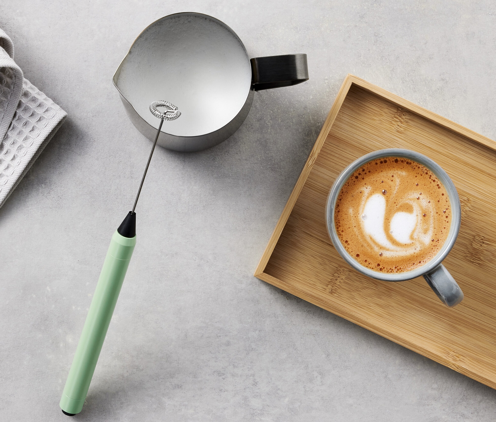 Serviette grise, pot, mousseur à lait et café au lait sur plateau en bois.