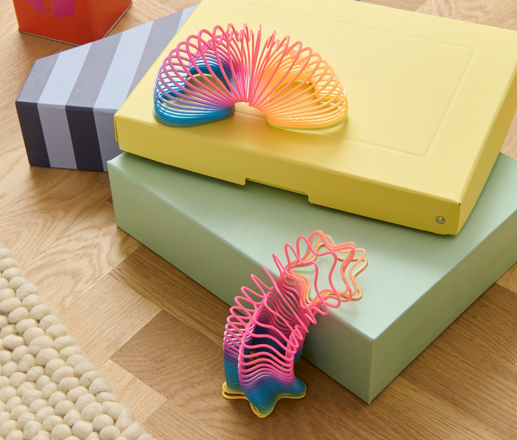 Deux jouets en ressort sont posés l'un sur l'autre sur trois boîtes et à côté d'une boîte en fer rouge sur un plancher en bois.