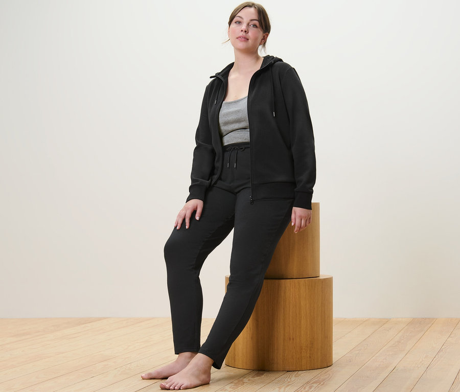 Femme assise sur des cylindres en bois portant une jaquette en sweat noire, un pantalon en sweat noir et un top d'un ensemble de 2.
