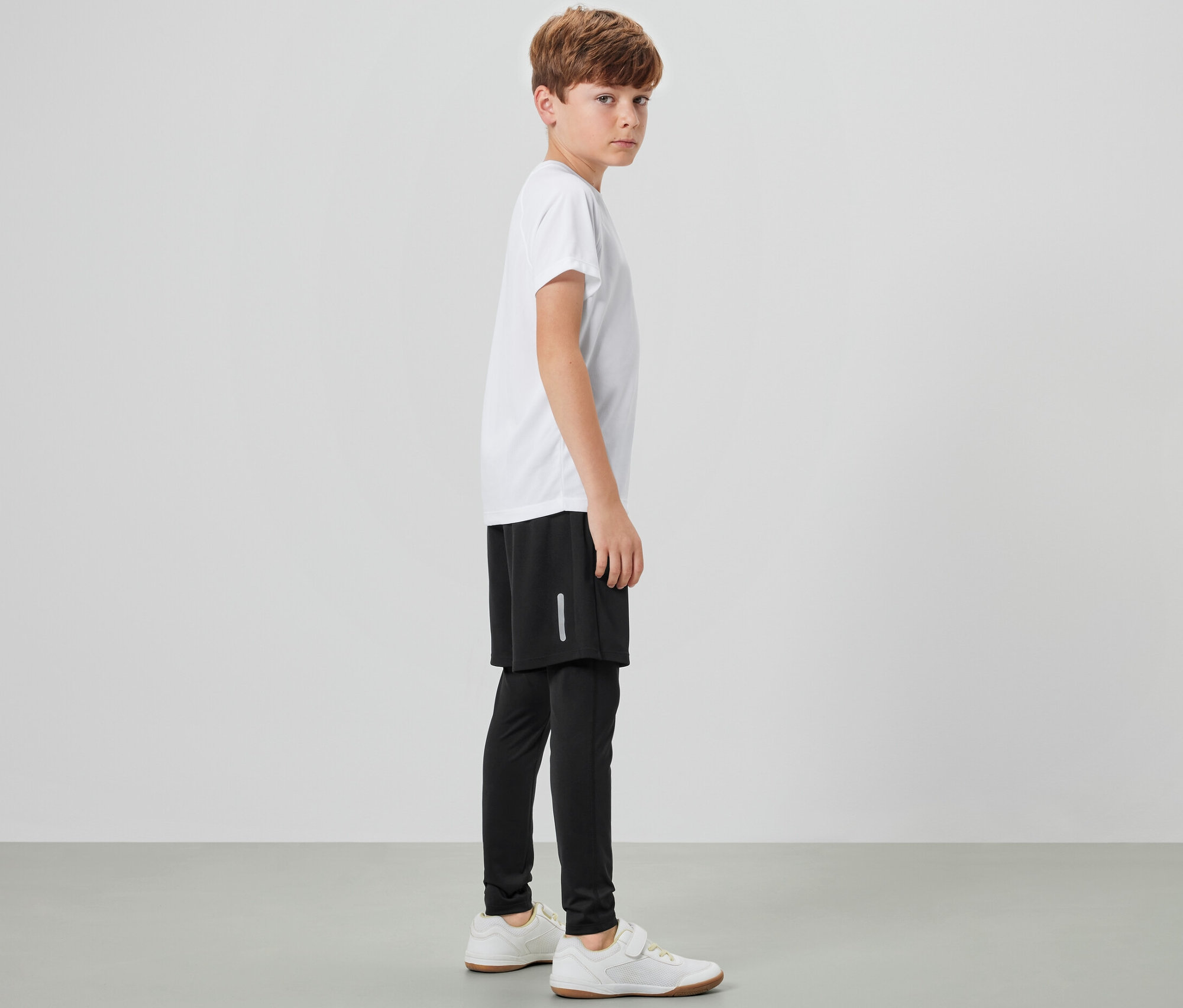 Un garçon se tient de côté et porte un tight de sport pour enfant avec short, un t-shirt blanc et des baskets.